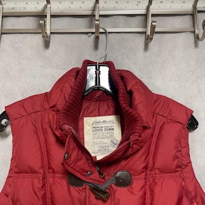 May include: A red Eddie Bauer goose down vest with a high collar and toggle closures. The vest is hanging on a black hanger. The vest has a quilted design and a label that reads "Premium Quality Goose Down."