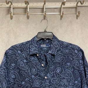 Puede incluir: Camisa abotonada azul marino con un estampado de cachemira en tonos azules y blancos. La camisa está hecha de una tela ligera y presenta un cuello clásico y puños abotonados. La etiqueta Polo by Ralph Lauren es visible.