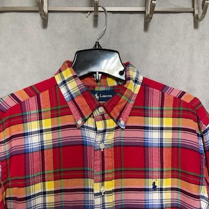 Op de afbeelding: Een rood, geel en blauw geruit button-down overhemd met een button-down kraag. Het overhemd heeft een klein geborduurd logo van een persoon op een paard. Het overhemd hangt aan een zwarte hanger.