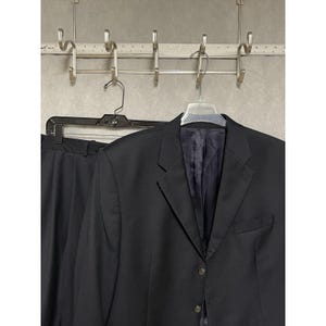 May include: A black suit jacket and matching trousers hang on a hanger. The jacket has three buttons and a notched lapel. The suit is made of a dark fabric and is displayed against a neutral background.