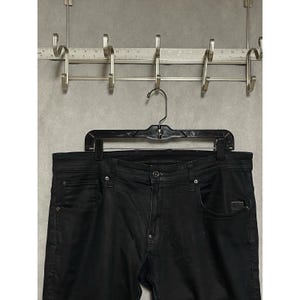 Könnte beinhalten: Schwarze Jeans aus Denim mit Knopfleiste, Fünf-Taschen-Design mit Metallnieten und einem kleinen rechteckigen Markenetikett an der vorderen rechten Tasche. Ein Metalllineal ist über dem Kleiderbügel sichtbar und zeigt Maße in Zentimetern.
