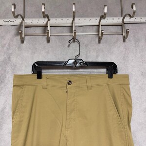 May include: Khaki-coloured trousers hanging on a black hanger. The trousers have a button closure and pockets. The background features a metal rack with hooks and a measuring ruler.