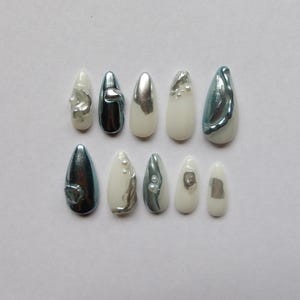 Op de afbeelding: Een set van tien kunstnagels in verschillende vormen en ontwerpen. De nagels hebben een combinatie van witte, zilveren en blauwgroene kleuren. Sommige nagels hebben parelaccenten of metallic afwerkingen, andere zijn effen van kleur.