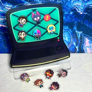 Puede incluir: Una pantalla acrílica transparente con un diseño de televisión retro negro con una pantalla verde y pines de personajes de dibujos animados. Los pines incluyen varios personajes con diferentes colores de cabello y atuendos. Se muestran pines adicionales debajo.