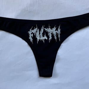 May include: Black thong underwear with the word "FILTH" in a silver, gothic-style font across the front. The underwear has a low-rise waist and a thong back.