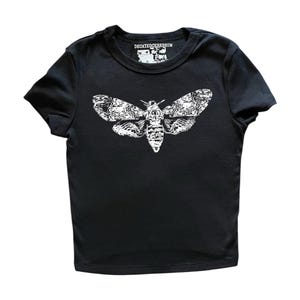 May include: A black short-sleeved t-shirt featuring a white graphic print of a moth on the chest. The inner neck label displays the text DECAYEDCEREBRUM in white lettering. The garment has a crew neckline and a straight hemline design.