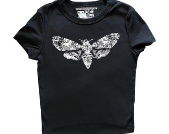 May include: A black short-sleeved t-shirt featuring a white graphic print of a moth on the chest. The inner neck label displays the text DECAYEDCEREBRUM in white lettering. The garment has a crew neckline and a straight hemline design.