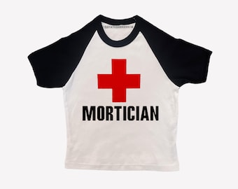 May include: A white raglan t-shirt with black sleeves and a black collar. The front features a red cross symbol above the word MORTICIAN in bold black capital letters. The inner neck label displays the text DECAYEDCEREBRUM in a black font.