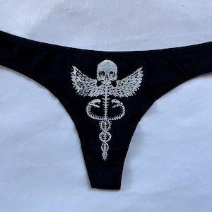 May include: Black thong underwear featuring a silver-colored graphic of a skull with wings above a caduceus symbol. The underwear is made of a soft, stretchy material.