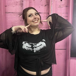 May include: Black top with a white graphic print and sheer sleeves. The top is tied at the waist. The background is a pink wooden wall.