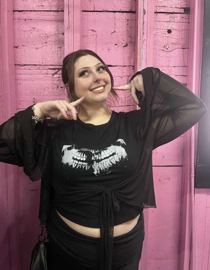 May include: Black top with a white graphic print and sheer sleeves. The top is tied at the waist. The background is a pink wooden wall.