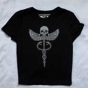 May include: A black short-sleeve t-shirt featuring a white graphic print of a winged skull atop a skeletal caduceus symbol. The inner neck label displays the text DECAYEDCEREBRUM in white lettering against a black background on the garment.