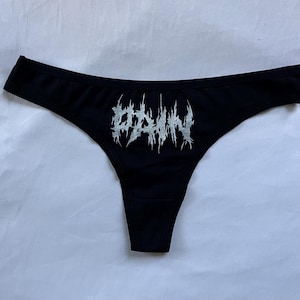 May include: Black thong underwear with a silver, stylized text graphic that reads "ODDIN". The underwear is made of a soft, stretchy material.