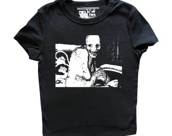 May include: A black short-sleeve t-shirt featuring a high-contrast white graphic print of a skeletal figure with a wide, toothy grin. The interior neck label displays the text DECAYEDCEREBRUM in white lettering against a black background.