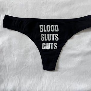 May include: Black thong underwear with the words "BLOOD SLUTS GUTS" printed in white. The underwear is displayed on a white surface. The text is in a bold, sans-serif font.