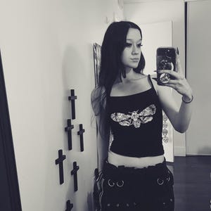 May include: Black crop top with a moth graphic. The top has thin straps and is paired with black pants featuring multiple silver rings and pockets. The image is in black and white.