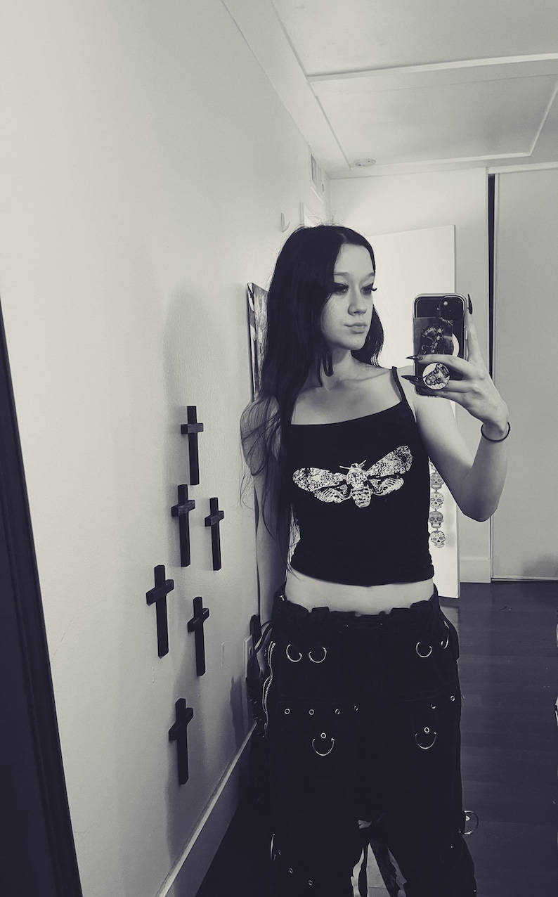 May include: Black crop top with a moth graphic. The top has thin straps and is paired with black pants featuring multiple silver rings and pockets. The image is in black and white.