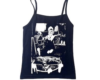 May include: A black spaghetti strap tank top featuring a high-contrast white graphic print. The design depicts a person with an updo hairstyle reading a book near candles and a dollhouse, with a doll lying inside a circular shape at the bottom.