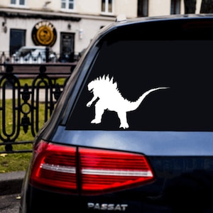 以下が含まれることがあります： 青い車のリアウィンドウに、ゴジラのシルエットの白いビニールステッカーが貼られています。車はフォルクスワーゲンパサートです。