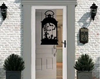 Puede incluir: Silueta negra de una linterna con una escena de Halloween espeluznante en su interior. La linterna está en una puerta blanca con un panel de vidrio.