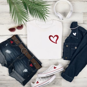 May include: A casual outfit laid out on a light wood-grain surface. It features a white t-shirt with a red heart, denim shorts, a denim jacket, white trainers with heart details, sunglasses, and headphones. The ensemble suggests a relaxed, everyday style.