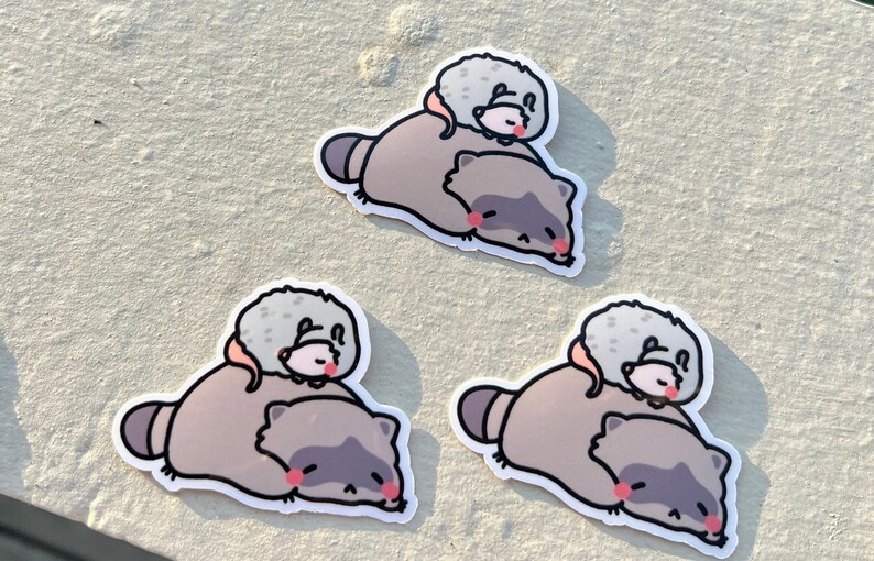 May include: Three stickers featuring a cartoon raccoon and a small gray animal. The raccoon is lying on its side with its eyes closed and the small animal is on top of the raccoon. The stickers are on a light gray surface.