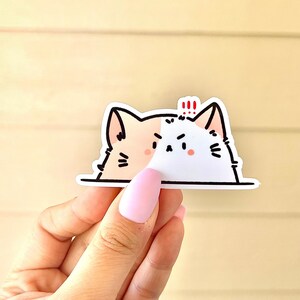 May include: A white and peach cat sticker with black eyes and a black nose. The cat is peeking over a white surface and has a red exclamation mark above its head.