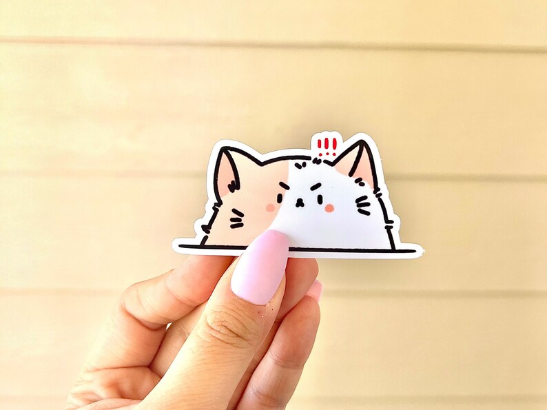 May include: A white and peach cat sticker with black eyes and a black nose. The cat is peeking over a white surface and has a red exclamation mark above its head.