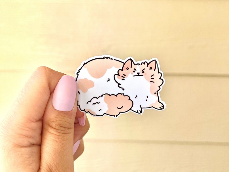 May include: A white and peach cat sticker with black outlines. The cat is sitting with its back to the camera and its tail curled around its body. The cat has a grumpy expression on its face.