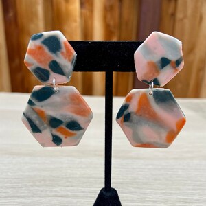 Könnte beinhalten: Ein Paar Hängeohrringe mit geometrischer Hexagonform. Die Ohrringe sind aus Polymer Clay gefertigt und haben ein marmoriertes Muster in den Farben Rosa, Orange, Schwarz und Weiß.