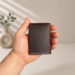 以下が含まれることがあります： 手に持たれた濃い茶色の革製財布。財布は長方形で角が丸く、底の端に沿ってステッチが見えます。革は使い古したような外観をしています。