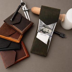 Puede incluir: Una colección de carteras de cuero hechas a mano en varios colores, incluyendo marrón, negro y verde. Una cartera está abierta, mostrando un billete de 100 dólares y una tarjeta de crédito. También se ven herramientas de artesanía, como un compás y un mazo.