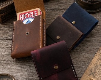 May include: A collection of handmade leather card cases in various colors: brown, navy, dark brown, and burgundy. One case is open, revealing a Bicycle playing card deck. The cases have a snap closure and white stitching. The background includes a compass and a vintage map.