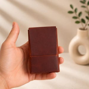 以下が含まれることがあります： 長方形の濃い茶色の革製財布。フラップ開閉式。手で持っており、豊かな色と質感を際立たせています。デザインはミニマルでクラシックで、高品質な職人技に焦点を当てていることを示唆しています。