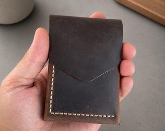 May include: A dark brown leather card holder featuring a pointed flap closure and visible light-colored stitching along the bottom and side edges. The leather has a matte, distressed finish with natural variations in the dark brown surface texture.