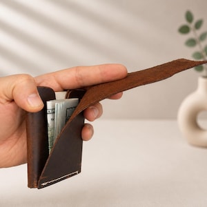 以下が含まれることがあります： 茶色の革製財布が部分的に開かれ、紙幣の束が見えています。財布は手で持たれており、そのミニマルなデザインと革の質感を強調しています。背景には、植物が入った小さなオフホワイトの花瓶があります。