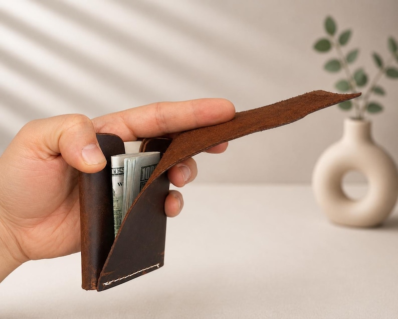 以下が含まれることがあります： 茶色の革製財布が部分的に開かれ、紙幣の束が見えています。財布は手で持たれており、そのミニマルなデザインと革の質感を強調しています。背景には、植物が入った小さなオフホワイトの花瓶があります。