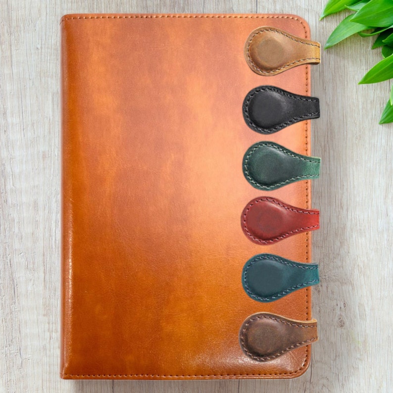 Puede incluir: Seis marcap&aacute;ginas magn&eacute;ticos de cuero en diferentes colores, incluyendo marr&oacute;n, negro, verde, rojo, verde azulado y marr&oacute;n, unidos a un diario encuadernado en cuero marr&oacute;n.