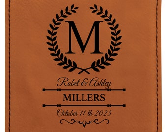 May include: Brown leather coaster with a black monogram design. The monogram features a wreath with the letter "M" in the center. The text "Robert & Ashley MILLERS October 11th 2023" is also included.