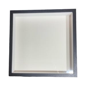 May include: Square shadow box frame with a dark gray wooden frame and a white interior. The frame has a recessed design, creating a three-dimensional effect. The frame is empty, ready for display.