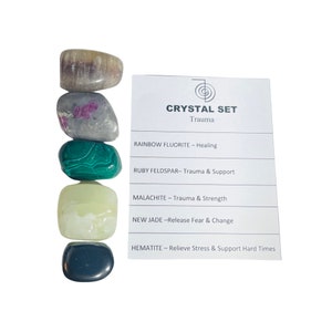 May include: A set of six tumbled crystals for healing trauma. The crystals are stacked on top of each other and include rainbow fluorite, ruby feldspar, malachite, new jade, and hematite. A white card with the names and healing properties of each crystal is shown to the right of the crystals.