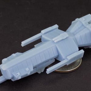May include: A light blue, miniature spaceship model with a detailed, angular design. The model features rectangular and cylindrical components, with wing-like extensions. The spaceship is placed next to a U.S. quarter for size comparison.