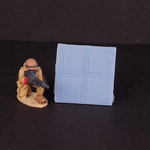 May include: A miniature plastic model of a soldier in a crouching position holding a rifle. The soldier is wearing a tan uniform and a helmet. The model is on a small base. The background is a blue and white tiled surface.