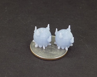 Pode incluir: Dois pequenos objetos esféricos de cor azul claro, impressos em 3D, com múltiplos espinhos. Os objetos estão sobre uma moeda de um quarto de dólar americano de prata.
