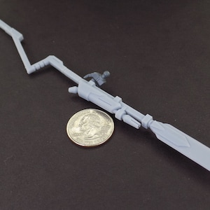 May include: A gray plastic model of a futuristic weapon, resembling a spear with a long, thin blade and a complex, articulated handle. The weapon is about 10 inches long and is lying on a dark gray surface next to a US quarter.