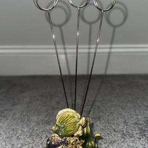 May include: A decorative metal photo holder with three wire loops. The base features a gold-colored design with two fish, coral, and faux gems. The fish are green and purple with jewel accents. The holder is suitable for displaying photos or notes.