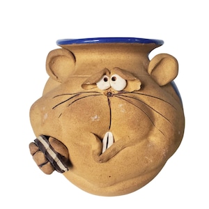 Op de afbeelding: Een grillige, handgemaakte keramische pot met een cartoongezicht. De pot is bruin met een blauwe rand en heeft grote ogen, snorharen en een tand. Een klein, bruin en wit object is aan de zijkant bevestigd.
