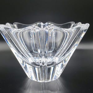 Puede incluir: Un cuenco de cristal transparente y facetado con un diseño de estrella. El cuenco tiene una base cuadrada que se ensancha hasta un borde festoneado. El cristal es transparente, lo que permite que la luz pase y se refleje en las facetas interiores.