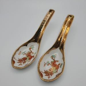 May include: Two ceramic Asian soup spoons. The spoons have a white interior with a red and gold dragon design. The rims and handles are gold. The spoons are approximately 6 inches long.
