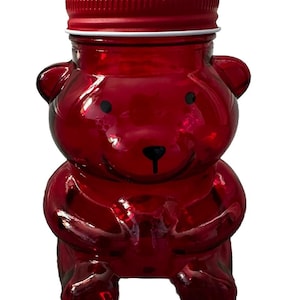 Pode incluir: Um frasco de vidro vermelho em forma de urso de pelúcia com uma tampa vermelha a condizer. O frasco tem olhos pretos e um nariz preto. O frasco foi concebido para parecer um urso a segurar as patas.